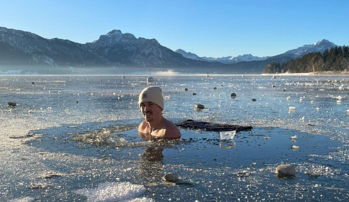 Thomas Kramer sitzt in einem freigehackten Eisloch in einem zugefrorenen See, dahinter verschneite Berge